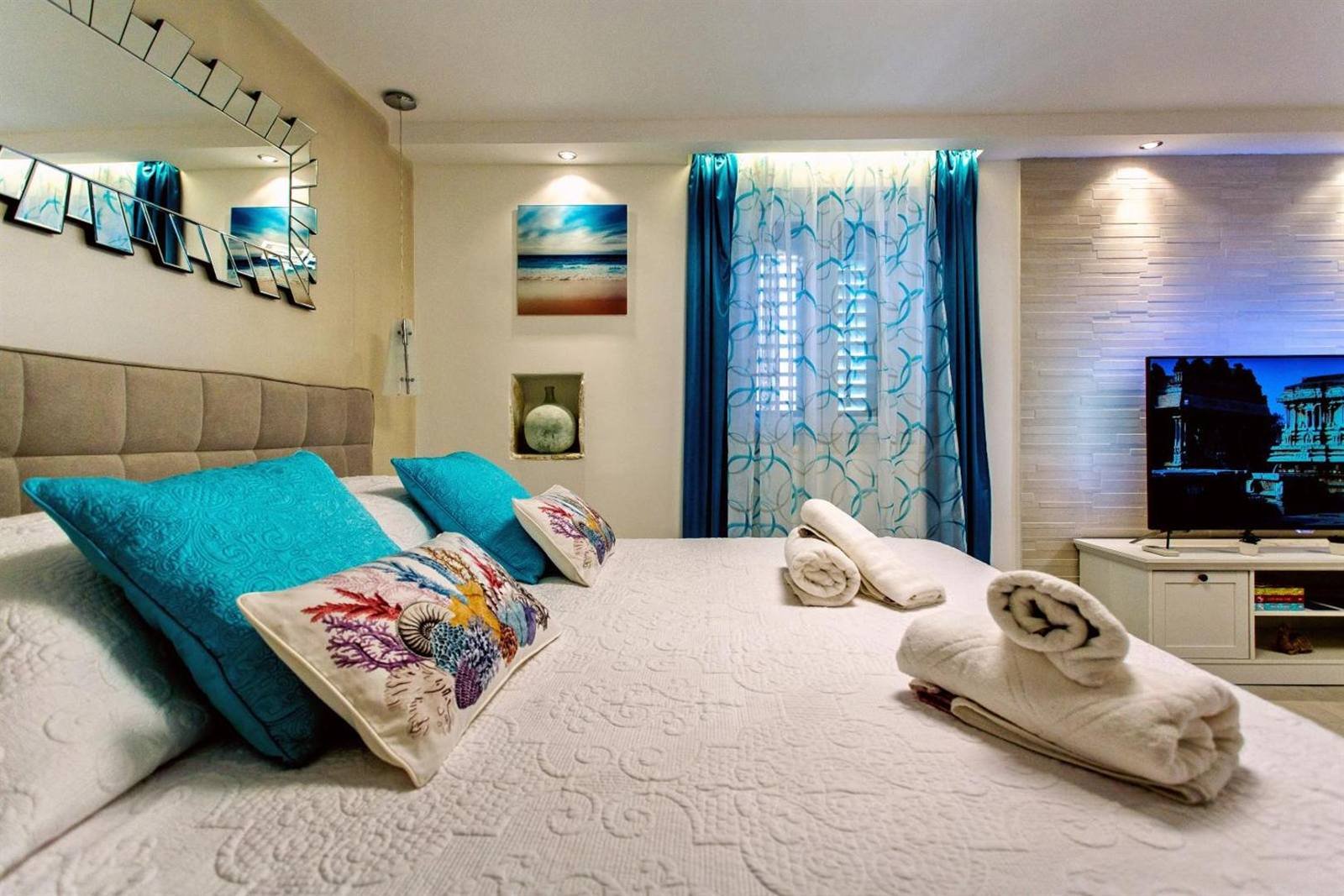 Double bed with turquoise cushions and folded towels, next to the TV and a window with blinds